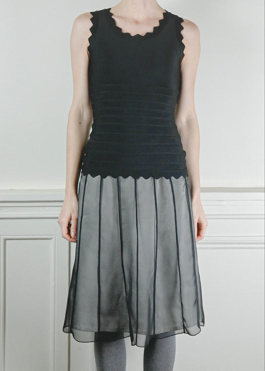 Person wearing a black top and gray skirt with tights and boots against a white paneled wall.