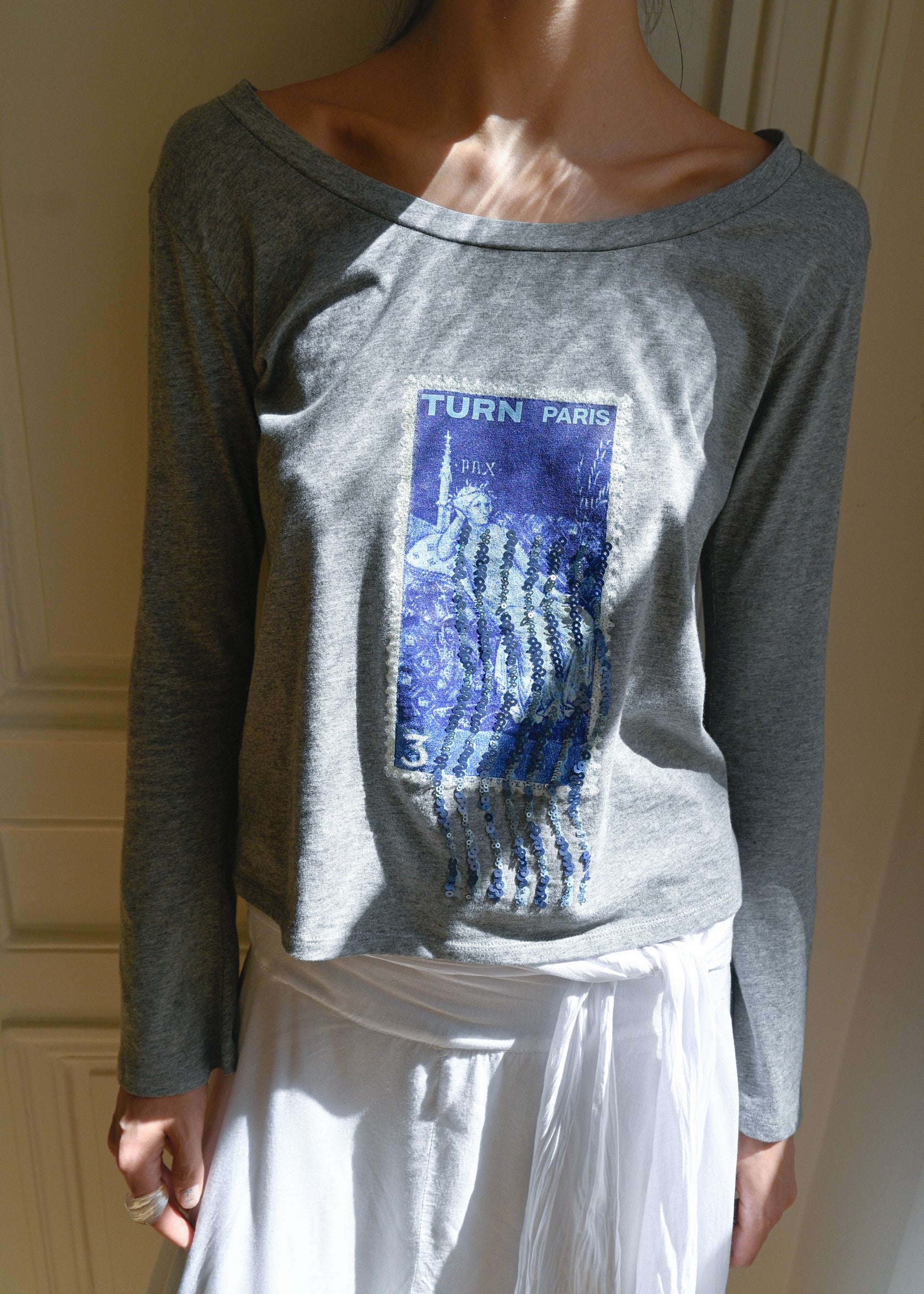 TURN PARIS long sleeve tee in soft grey, featuring a blue sequin-embroidered motif.

Inspired by the symbol of justice and the lucky number three.

Designed with a rounded open neckline that can be worn off the shoulder, and long flared sleeves.