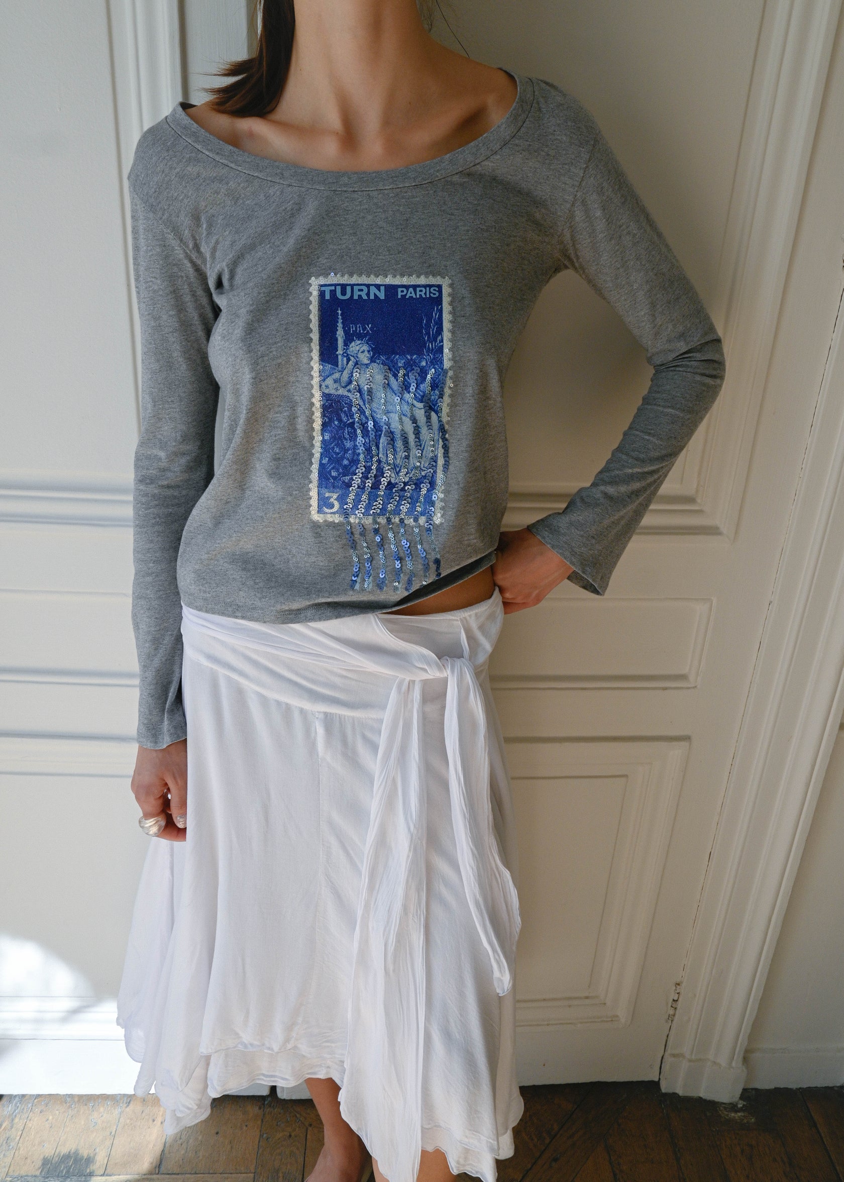 TURN PARIS long sleeve tee in soft grey, featuring a blue sequin-embroidered motif.

Inspired by the symbol of justice and the lucky number three.

Designed with a rounded open neckline that can be worn off the shoulder, and long flared sleeves.
