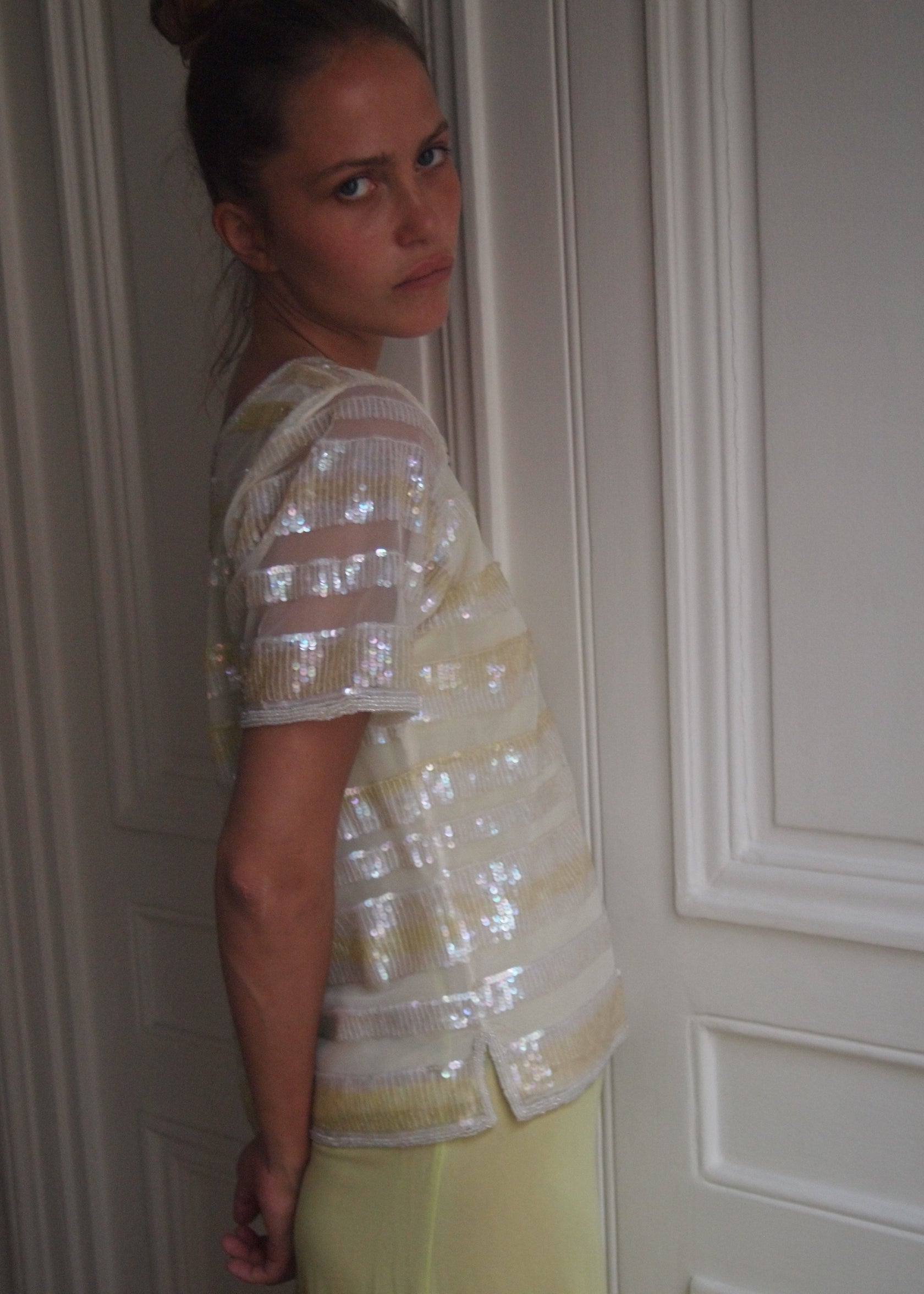 A woman wearing a sheer, short-sleeved top with horizontal metallic silver sequin stripes and pale yellow bottoms, standing in front of white paneled doors.
