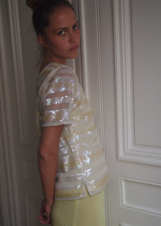 A woman wearing a sheer, short-sleeved top with horizontal metallic silver sequin stripes and pale yellow bottoms, standing in front of white paneled doors.