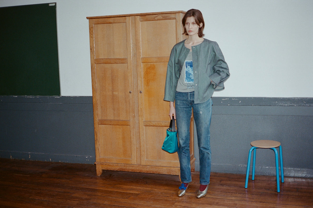 Model in a grey blue shirt and jeans with a teal bucket bag, standing by a wooden wardrobe in a classroom. TURN Paris FW26, Paire collection