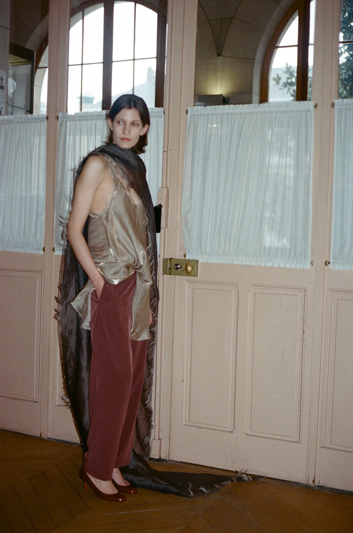 Model in a gold silky top and burgundy trousers with a long sheer scarf, by tall school doors. TURN Paris FW26, Paire collection