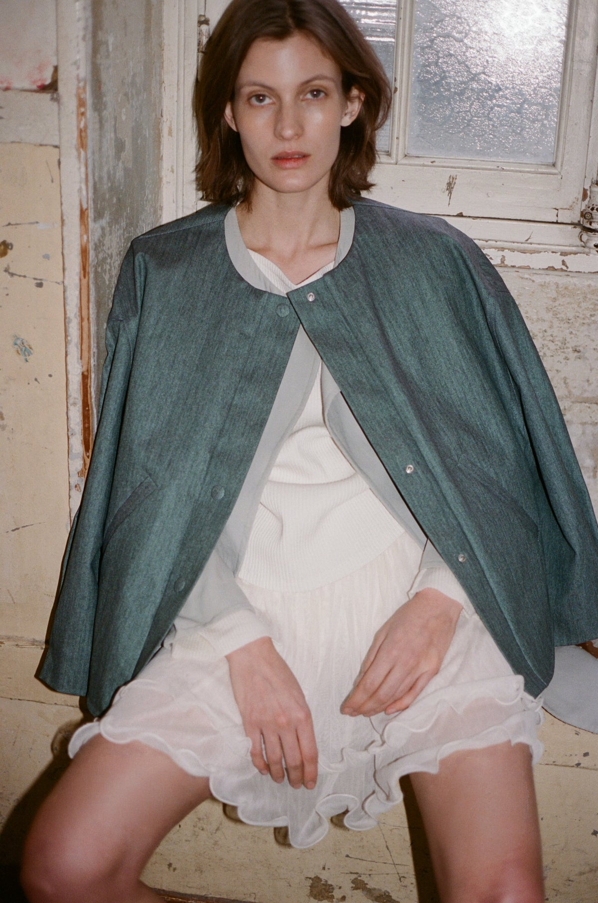 Model in an oversized green shirt over a white lace slip dress, seated by a window. TURN Paris FW26, Paire collection