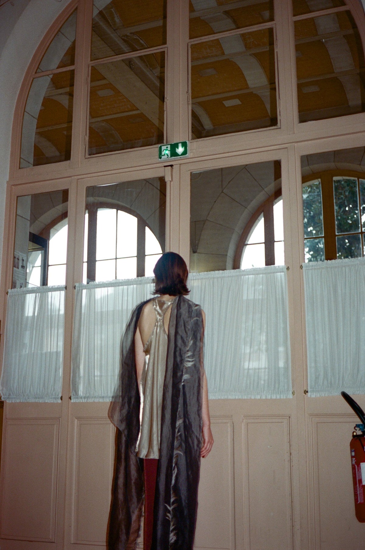 Model seen from behind wearing a long printed scarf in a school hall with arched windows. TURN Paris FW26, Paire collection