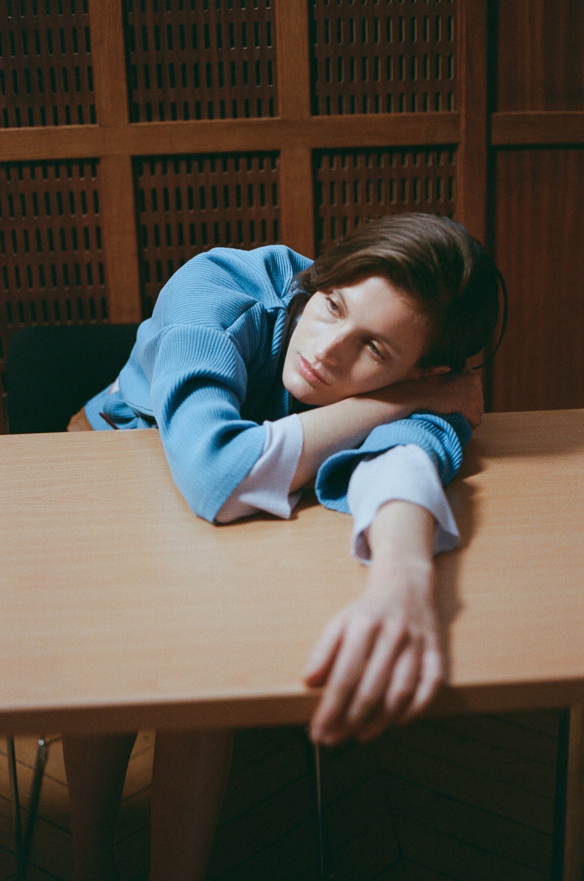 Model in a blue sweater resting her head on a wooden school desk. TURN Paris FW26, Paire collection