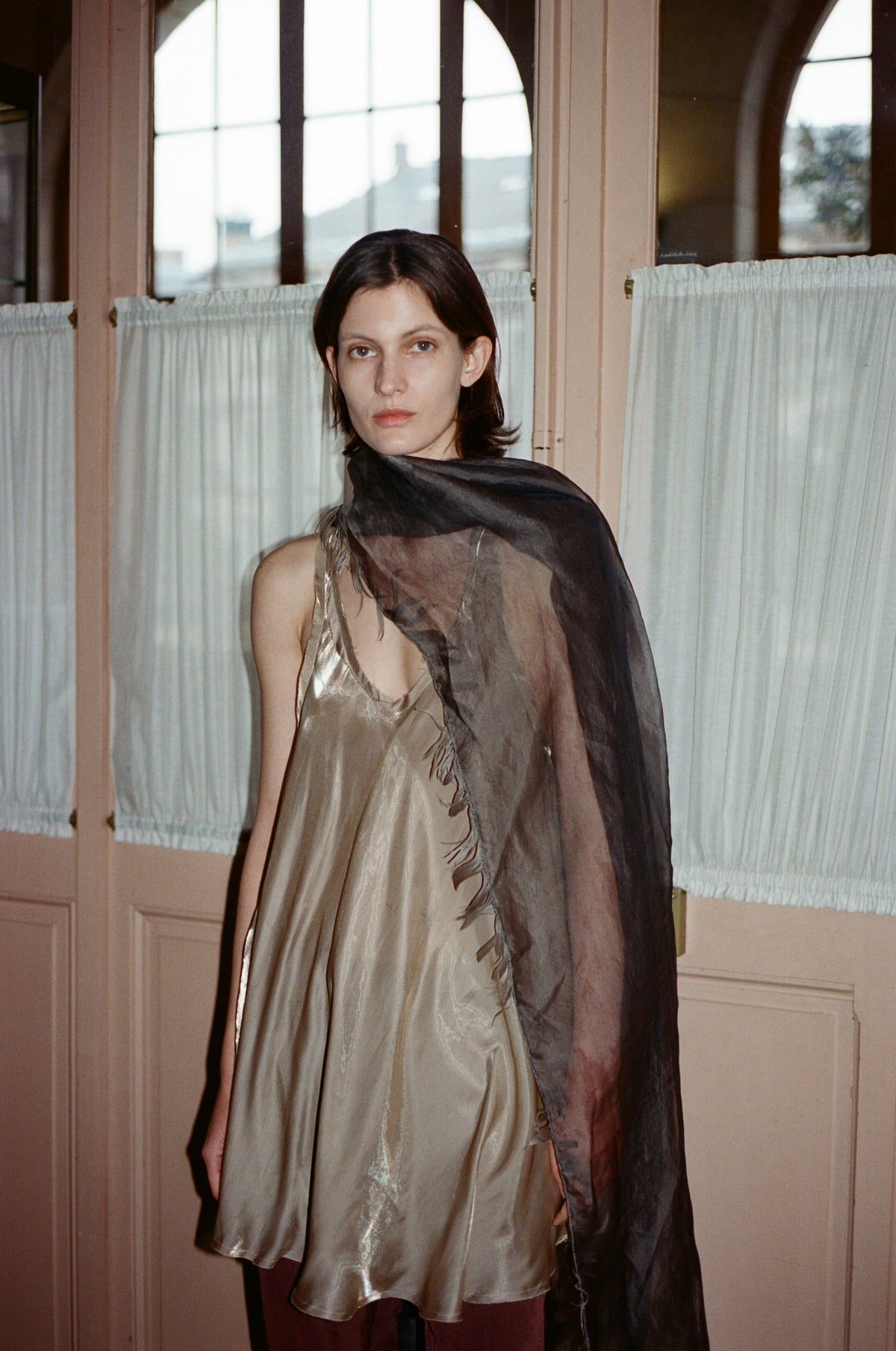 Model in a gold satin racer back tunique with a draped scarf, by school doors. TURN Paris FW26, Paire collection