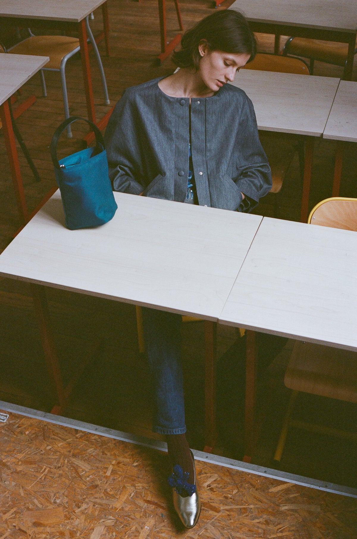 Model in a grey blue shirt seated at a classroom table with a teal bucket bag. TURN Paris FW26, Paire collection